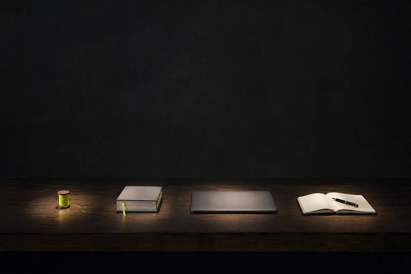 Quatro objetos sobre uma mesa escura: um carretel de fio verde-limão, páginas encadernadas, um laptop fechado e um caderno aberto.