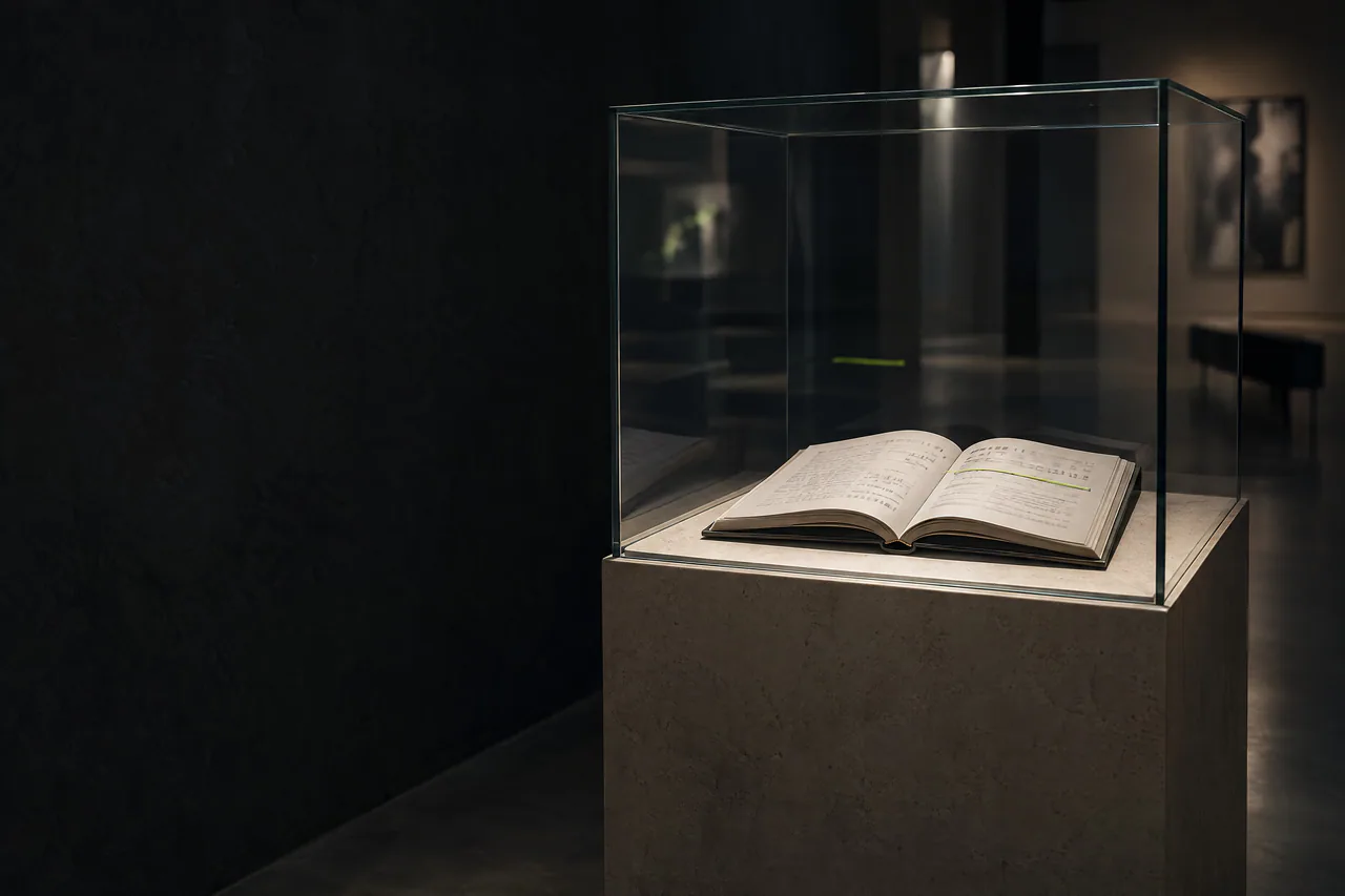 Um documento aberto dentro de uma vitrine de vidro em uma galeria escura, com uma linha marcada por um fio verde-limão.